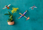 The Studio ROOF Robin Plane, a cardboard model plane with geometric patterns and bright colours, plus 2 other Studio ROOF planes, all hanging with a mottled teal wall behind. to the lower right there is a contrasting peach plant containing green plant wit