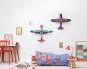 The Studio ROOF Deluxe Condition Glider Plane, a huge cardboard model plane with geometric patterns and bright colours, put together, on a white wall in a bedroom with the other Studio ROOF deluxe plane, above a Childs bed. a clock and picture are to the 