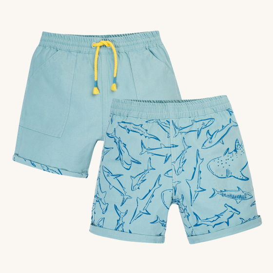 Frugi Children Organic Cotton Rocky Reversible Shorts - Stingray / Jawsome. A plain pale blue short with yellow drawstring on one side and a Jawsoms shark outline print on the reverse, on a cream background