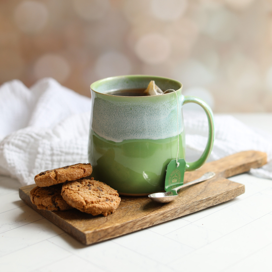 Glosters - Ceramic handmade mug in Pea Green. The mug has a tea bag inside and sits on a biscuit board. 