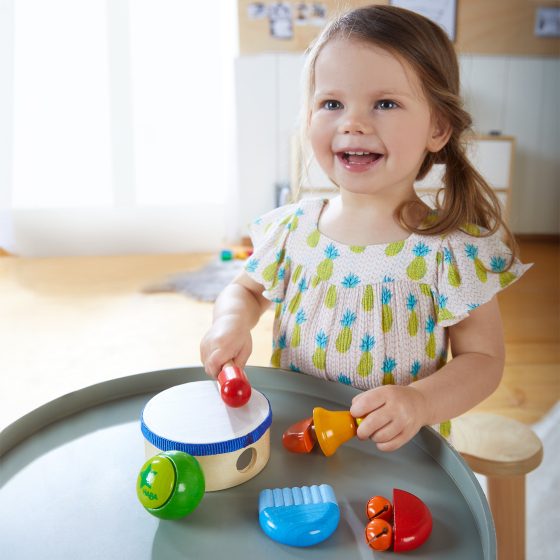 Haba Musical Instruments - Sounds Set. 6 musical instruments include a drum and drum stick, a shaker, a bell chime, a bell rattle and a guiro, with a child playing happily with the instruments