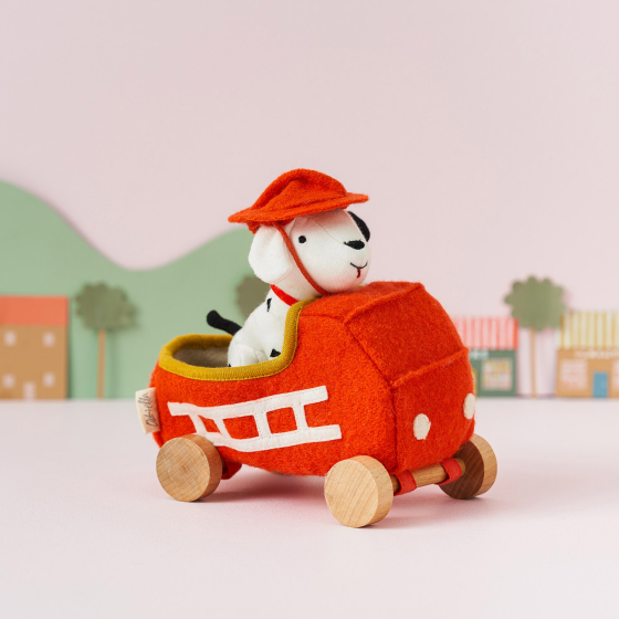 Olli Ella Holdie Dog-go Chief in a red felt fire truck with wooden wheels, wearing a red fire fighter helmet