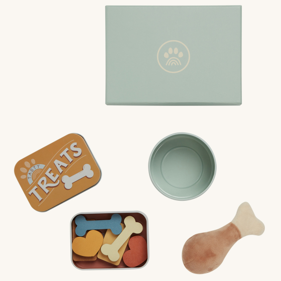 The Olli Ella Dinkum Dog Goodies Set is a set of play toys that can be used as accessories for your Dinkum Dog. This set contains a box with a small tin reading "Treats", a toy dog bowl, and a plush toy chicken bone, all in a pale teal box.