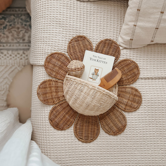 The Olli Ella Rattan Daisy Wall Basket in Natural Rattan on a bed, holding a small book, baby rattle and comb