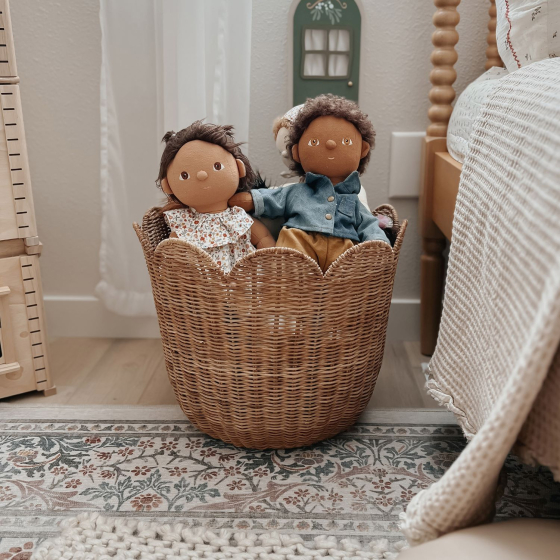 The Olli Ella Rattan Tulip Storage Basket in Natural, on a rug in a bedroom, holding a selection of Dinkum Dolls.