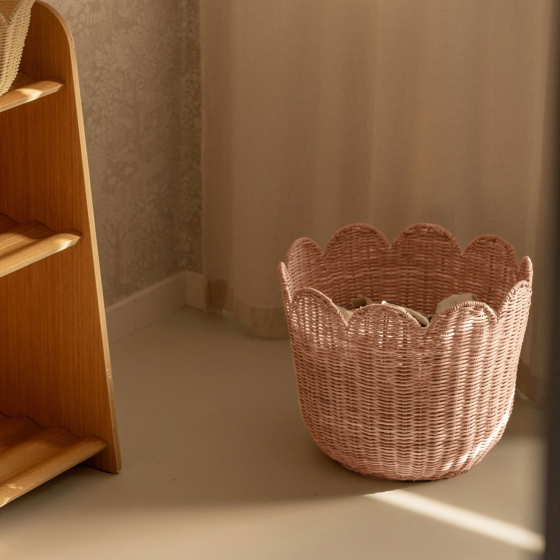 The Olli Ella Rattan Tulip Storage Basket in Seashell Pink on the floor in the corner of a room with a curtain in the background.