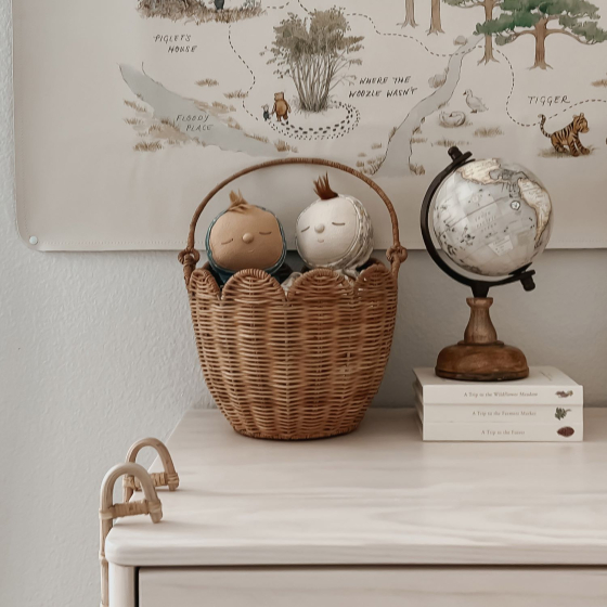 The Olli Ella Rattan Tulip Carry Basket - Natural holding two Dozy Dinkums on a chest of draws next to a globe, with a map on the wall behind.