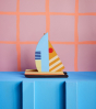 The Studio ROOF Halfmoon Catamaran, a cardboard model boat with geometric patterns and bright colours, put together, stood on a blue surface,  a pink background with orange check stripes 