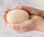 A bar of Kokoso Natural Organic Baby Soap, being held by a pair of hands with manicured nails. Marble effect background out of focus. 