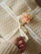 A fabric, light pink flower has been placed in the bow of the Avery Row branded ribbon. The blanket is a light beige colour, and the ribbon is being held between an adults fingers