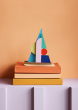 The Studio ROOF Eclipse Catamaran, a cardboard model boat with geometric patterns and bright colours, put together, with light orange wall background, on a stack of 3 books