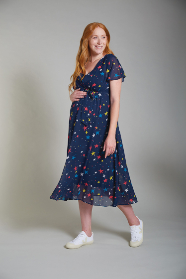 Smiling pregnant woman wearing the Frugi Isobel northern light printed wrap dress with white trainers and a grey background